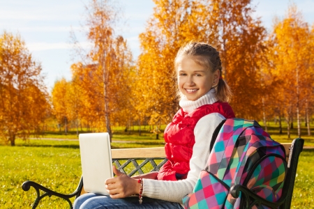 Happy blond 11 years old girl with amazing smile sitting on the bench with laptop turning back doing homework outside in the autumn park on sunny dayの写真素材