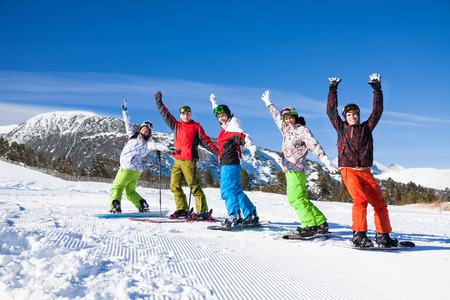 One skier and 4 snowboarders standing in a row and lifting hands up in the air and making fun on the mountains backgroundの写真素材