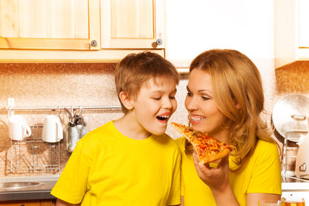 Cheerful mother feeding her smiling son with one hand with piece of pizza in the kitchenの写真素材