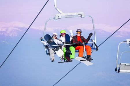 Three excited young people with snowboarders sitting on ropeway on the mountains backgroundの写真素材