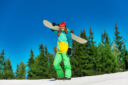 Smiling girl in ski mask standing and holding snowboard in winter with fir trees on the backgroundの写真素材