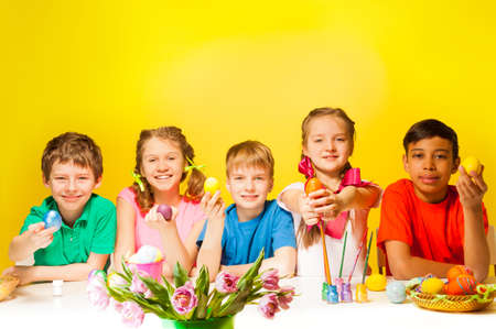 Five children holding coloured Easter eggs at the white desk with tulips on the yellow backgroundの写真素材