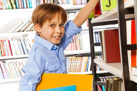 Happy schoolboy searching books and holding exercise books on the bookshelf in library