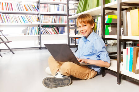Happy boy sitting with laptop on the floor in library