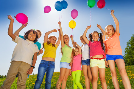 Children standing in half round with arms up to flying balloons in green fieldの写真素材