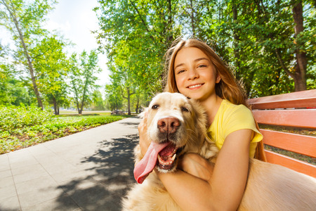 Portrait of teenage girl with big smile hugging cute golden retriever dog sitting on the bench in summer parkの写真素材