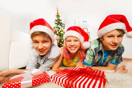 Three happy teen kids laying on the floor in the living room near Christmas tree in Santa caps smiling and laughing with red presentsの写真素材