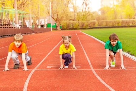 Three kids in colorful uniforms stand with bended knee ready to run the marathon together on the stadiumの写真素材