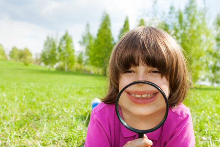 Funny boy smiles through magnifier laying alone on the green grass during beautiful summer day in parkの写真素材