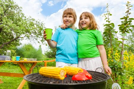 Close-up of small sisters standing near BBQ and grilling corn with paprika in the garden outside during summer dayの写真素材