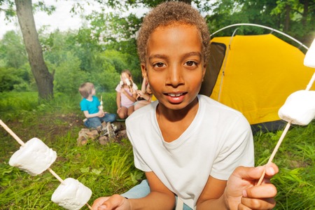 Portrait of African boy who holds sticks with marshmallows during camping in the forest near tentの写真素材