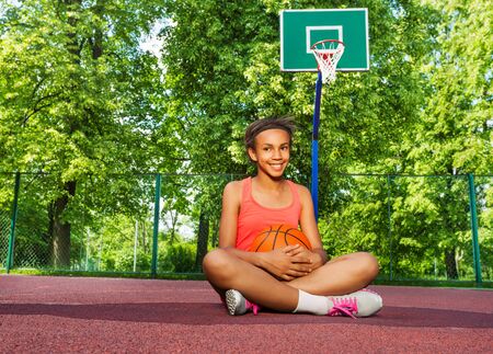 Smiling African girl sitting on the playground and holding the ball during sunny summer day aloneの写真素材