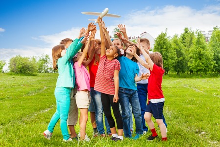 Group of kids reach after big white airplane toy with arms standing close in the field during summer dayの写真素材