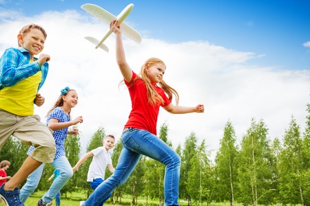 View from below of girl holding big white airplane toy and children behind running in the field during summer dayの写真素材