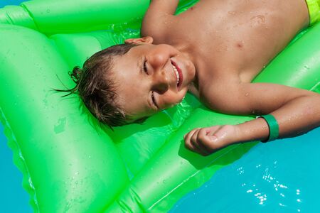 Close-up of happy boy relaxing on green inflatable mattress in swimming pool during summerの写真素材