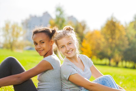 African and blond girl sitting together in park during sunny autumn dayの写真素材
