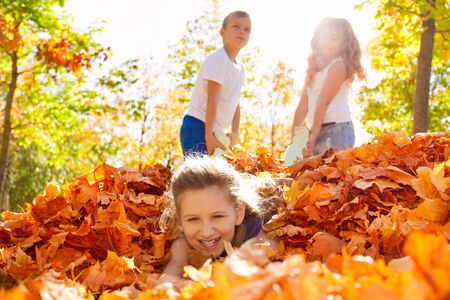 Children having fun dragging girl laying on the ground with leaves in forest during beautiful autumn sunny dayの写真素材
