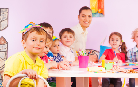 Kindergarten class with kids boys and girls and teacher sit by the table smiling and looking at cameraの写真素材