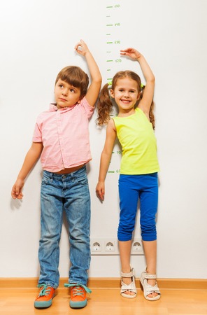 Two kids boy and little girl stand by the wall and show their height with hand above headの写真素材