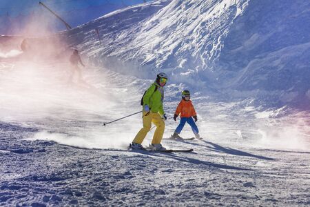 Mom and child ski down the piste teaching how to go down with sunlight lit through snow mistの写真素材