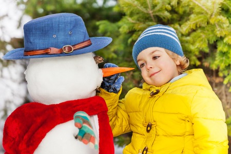 Little blond boy put carrot nose to snowman in the winter forestの写真素材