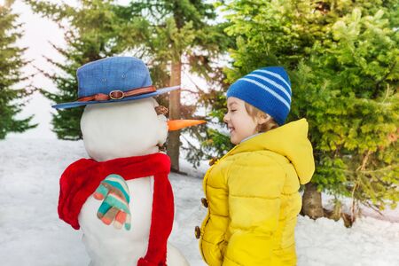 Little boy in yellow coat stand looking on the dressed in hat and scarf snowman he just madeの写真素材