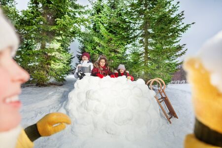 Picture of smiling girl throwing snowball at her friends hiding behind big snow tower, winter...