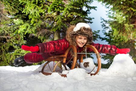 Cute funny girl in colorful winter clothes having fun on wooden snow sledge at the forestの写真素材