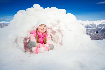Happy girl in pink clothes laying at the snow hole against beautiful mountainsの写真素材