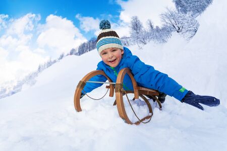 Little boy slide down on sledge smiling and looking at camera stretching hands