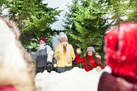 Happy friends, girls and boy playing snowballs hiding behind snow tower in the winter forest