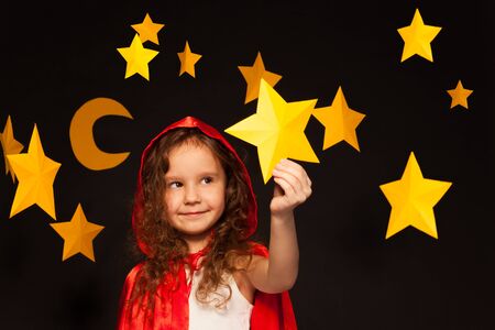 Little girl in red cape with hood holding big yellow paper star in her hand, standing against starry...