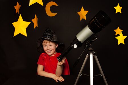 Happy five years old boy in sparkling cap and red cape with a telescope on black backgroundの写真素材