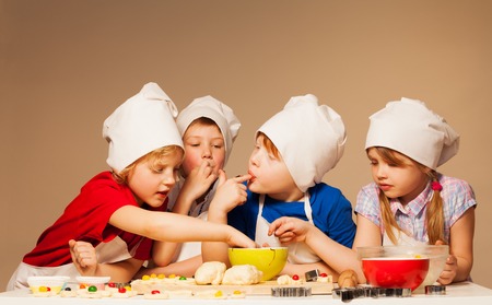 Four cute kids, boys and girl in baker's uniform tasting dough for handmade cookies, licking their fingersの写真素材