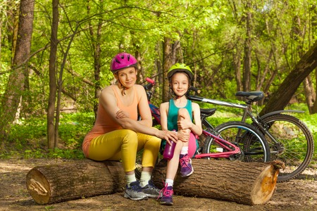 Cute little girl and her mom rest on logs in the spring sunny park, drinking water after cyclingの写真素材
