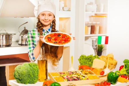 Cute young Italian girl in cook's uniform holding plate with tasty pizza in the kitchen full of kitchen-stuffの写真素材