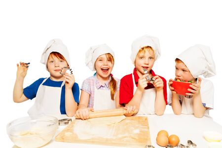Four young bakers, kids in aprons and toques standing in row, making dough for homemade cookies, isolated on whiteの写真素材