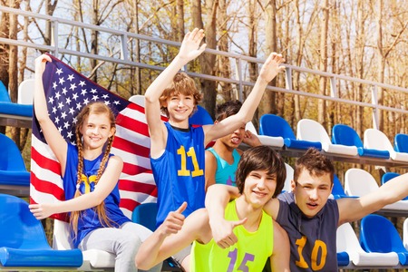 Happy teenage supporters celebrating the win of their sport team, sitting at tribune with American flagの写真素材