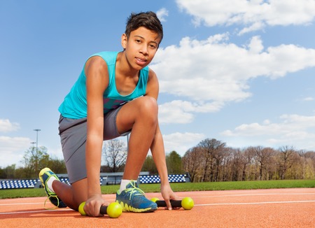 Teenage African sporty boy picking up two dumbbells, kneeling on track in the stadium