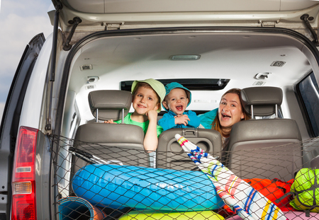 Cute young mother with her age-diverse sons peeking from behind a minivan seat, view from the boot full of luggageの写真素材