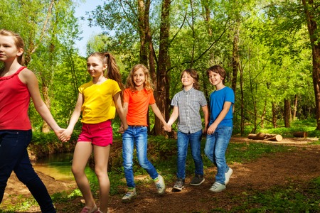 Portrait of five kids, ten years old boys and girls, walking in the green summer forest one after another holding handsの写真素材