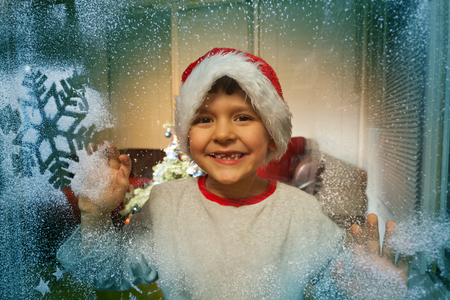 Boy in Santa hat behind frozen window with snowの写真素材