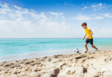 Portrait of cute boy in yellow t-shirt playing football on sandy sunny beachの写真素材
