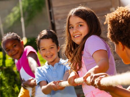 Rope pulling competition game with kids in a row in full action pull smile, focus on Caucasian long hair little girl, on summer dayの写真素材
