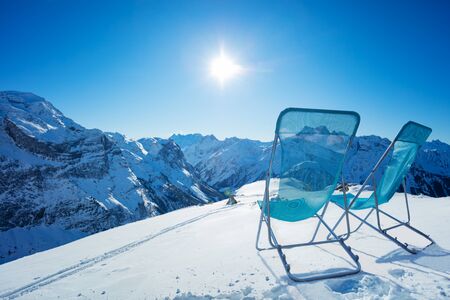 Two chaise longue chairs stand on the snow high in the mountains over snowy summit on sunny bright dayの写真素材