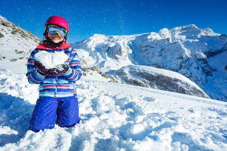 Smiling happy cute girl in ski outfit mask and helmet show love concept holding snow shape in heart standing on knees with mountains on backの写真素材
