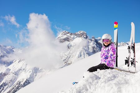 Happy girl in ski outfit, helmet and mask sit in the snow on mountain top look at cameraの写真素材