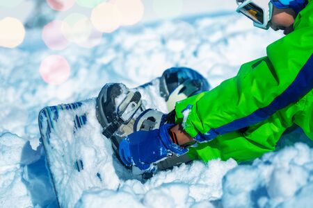 Close up of a boy attach snowboard to the legs sitting in snow about to start going downhill on ski resortの写真素材