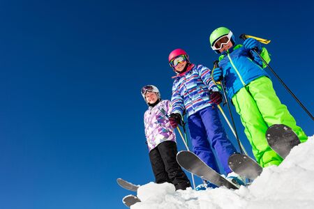 View from below of a group of three children stand on the mountain top in snow wearing ski colorful outfit over blue skyの写真素材