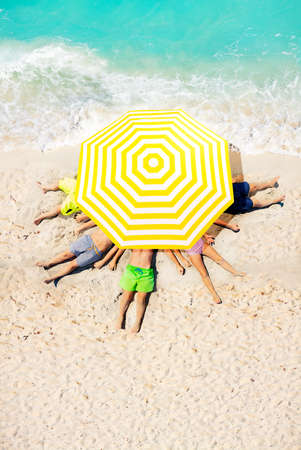 Group of kids lay on the beach sand under big sun umbrella view from above near sea wavesの写真素材